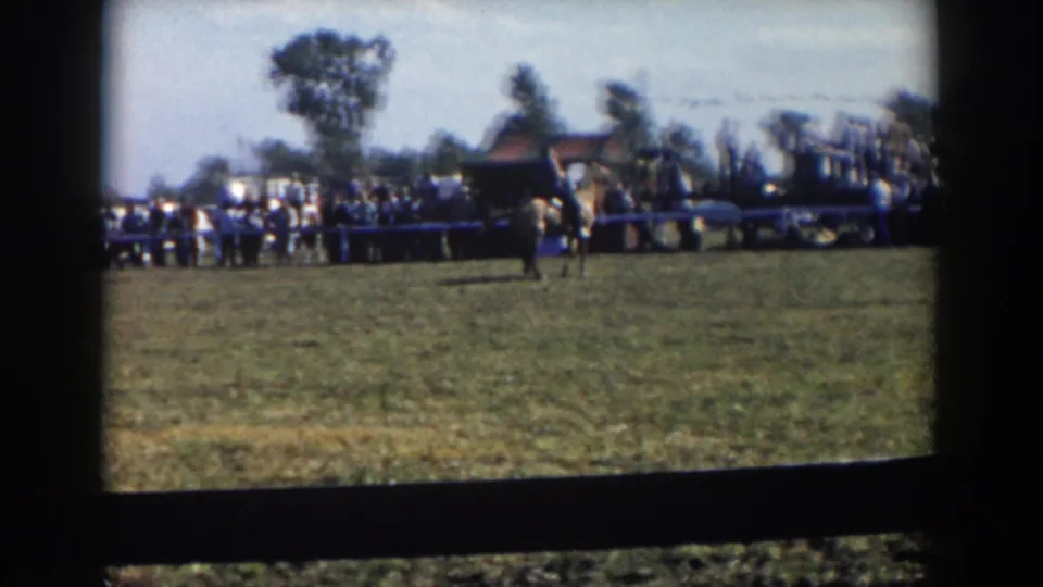 The camera holds a wider view of the Wheeling, Ohio fairground scene, showing spectators, parked cars, trees, and a figure on the field.