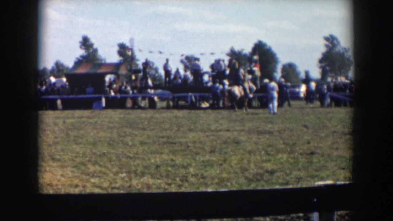 Horse riders and spectators stand near a fence at an outdoor race area.