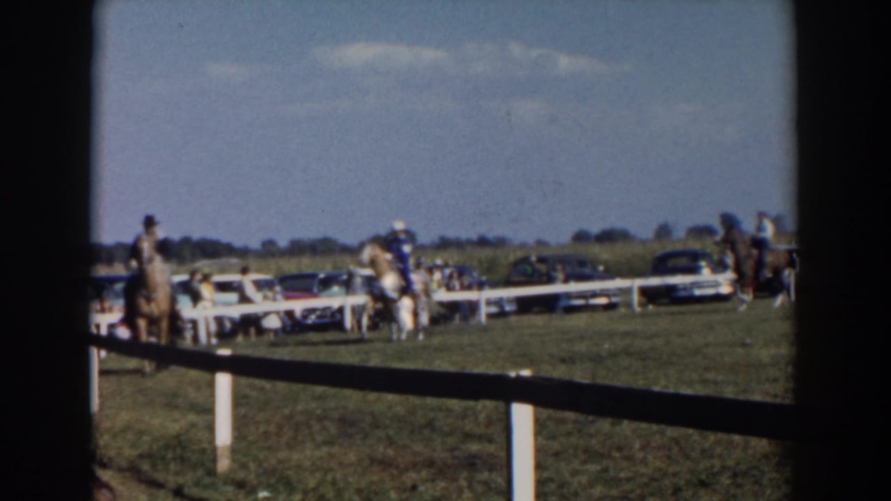 Horseback riders pass along the fenced grass ring with cars and spectators behind them.