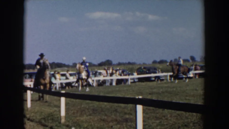 Home-movie footage frame shows multiple mounted riders in a fenced outdoor horse ring in Wheeling, Ohio, with spectators and cars beyond.