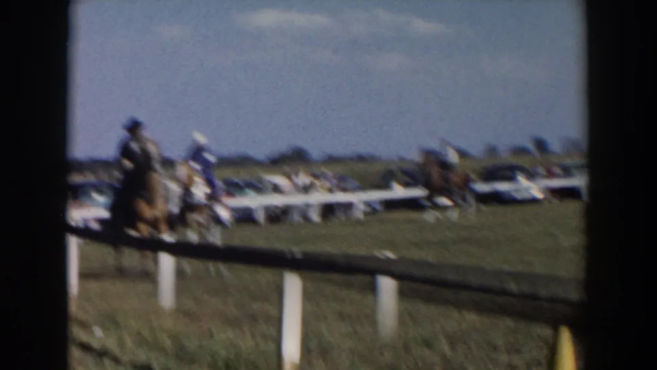 Motion-blurred 1957 frame from Wheeling, Ohio, shows horseback riders passing through a fenced grassy competition area with distant spectators.