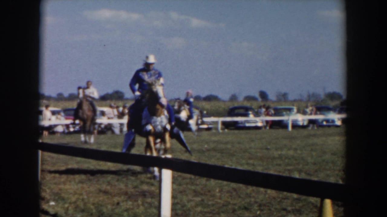 Rider in a white hat on a decorated horse crossing a grassy field near a white rail fence.