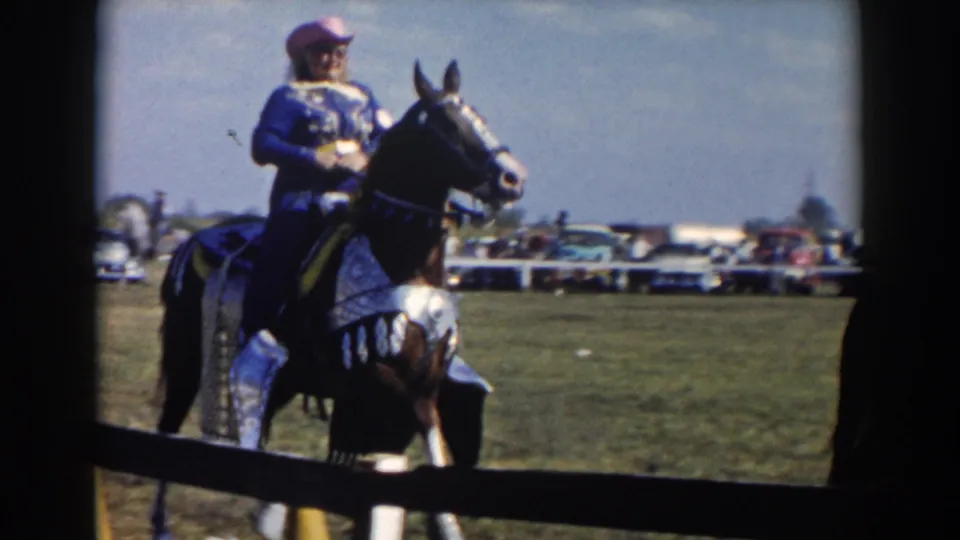 1957 home-movie frame identified as Wheeling, Ohio, showing a rider in blue on a decorated dark horse at a field event.