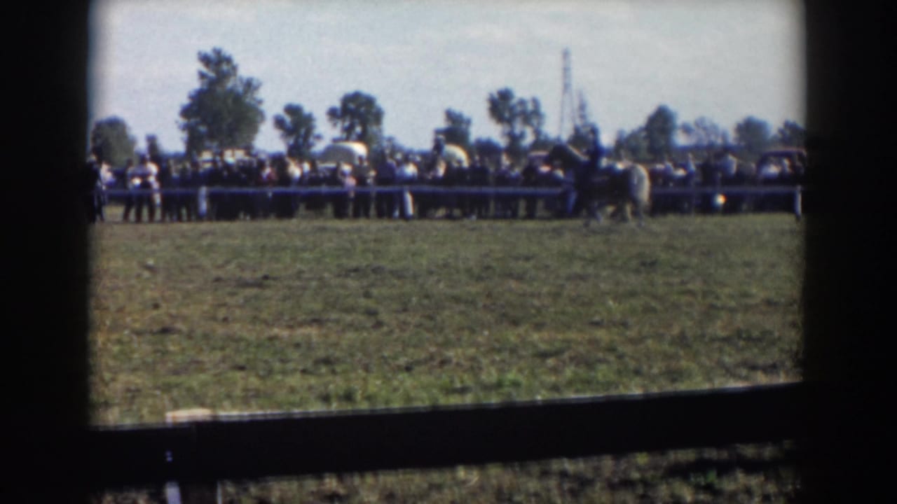 Wide field view with spectators behind a rail fence and riders in the distance.