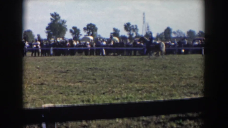 Wide 1957 event view with spectators along a fence and mounted riders crossing a grassy field in the distance.