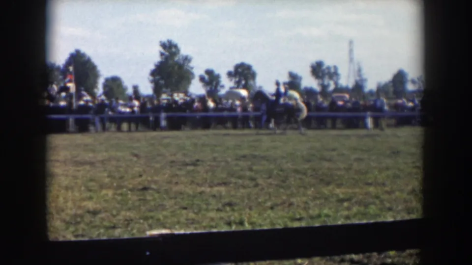 Home-movie frame of a horseback event with a rider at mid-distance and a line of spectators beyond the field.