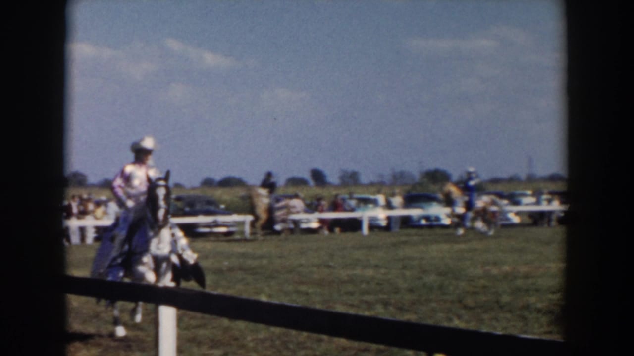 Horseback rider in a white hat moves near a rail fence with spectators and cars behind.