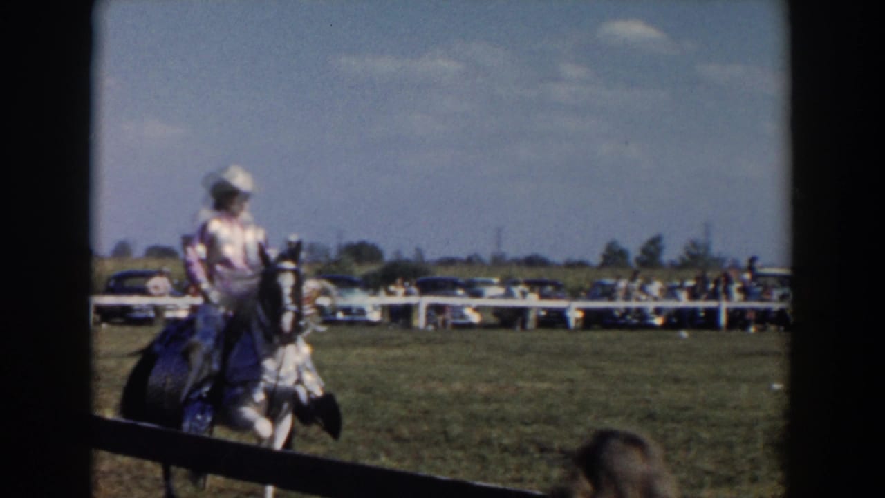 Rider on horseback crosses the field with cars and spectators beyond a white fence.
