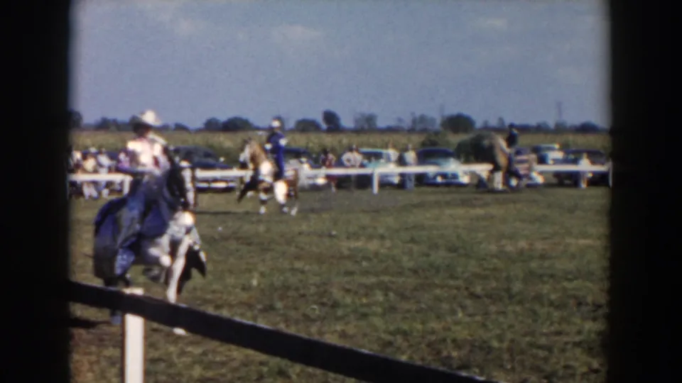 1957 Wheeling, Ohio frame with a horseback rider near the rail, additional riders, spectators, and parked cars across the field.