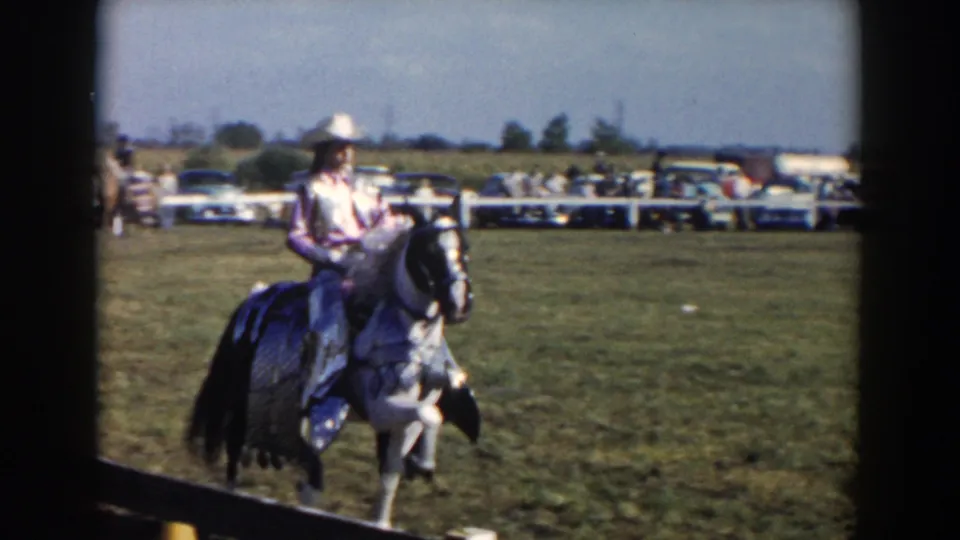 At a 1957 Wheeling, Ohio rodeo, a woman in western attire rides a black and white horse as spectators and cars line the background.
