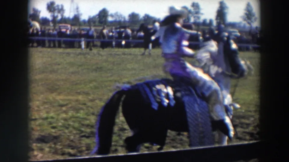 The black and white horse turns or passes close to camera, showing motion blur, western tack, grass, fence, and spectators beyond.