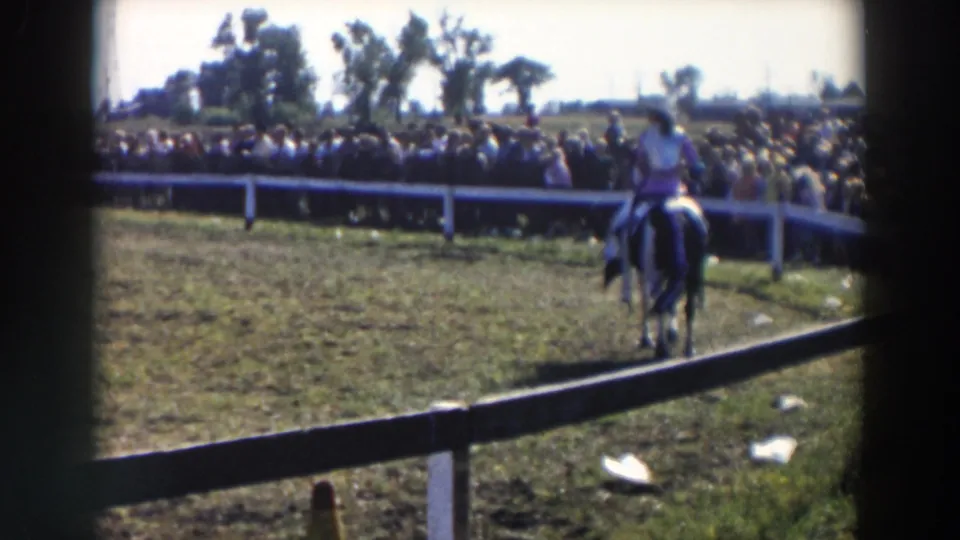 The woman rider heads away across the grassy Wheeling rodeo ring, with a long spectator line behind a white fence in bright daylight.