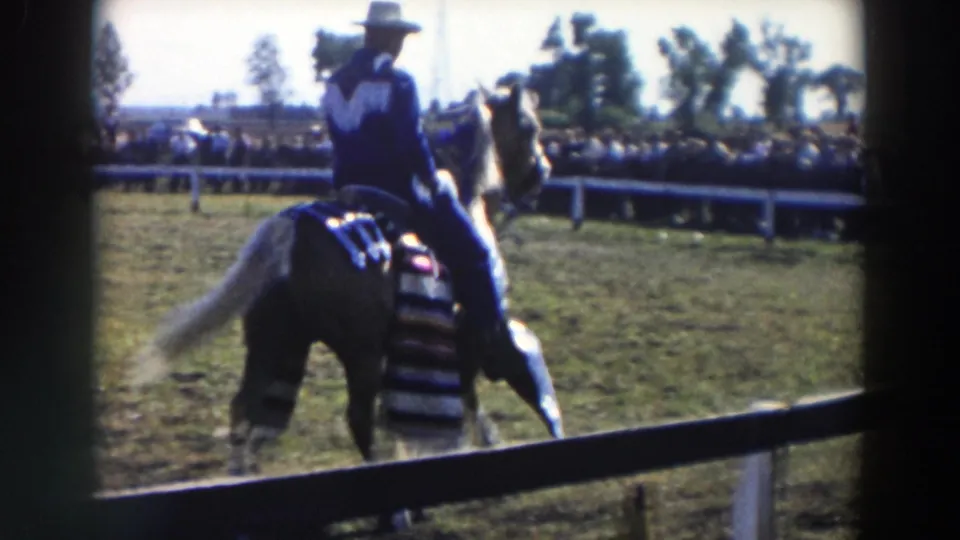 1957 8mm frame showing the horse and rider near a low arena rail, with a crowd watching from the far side of the field.