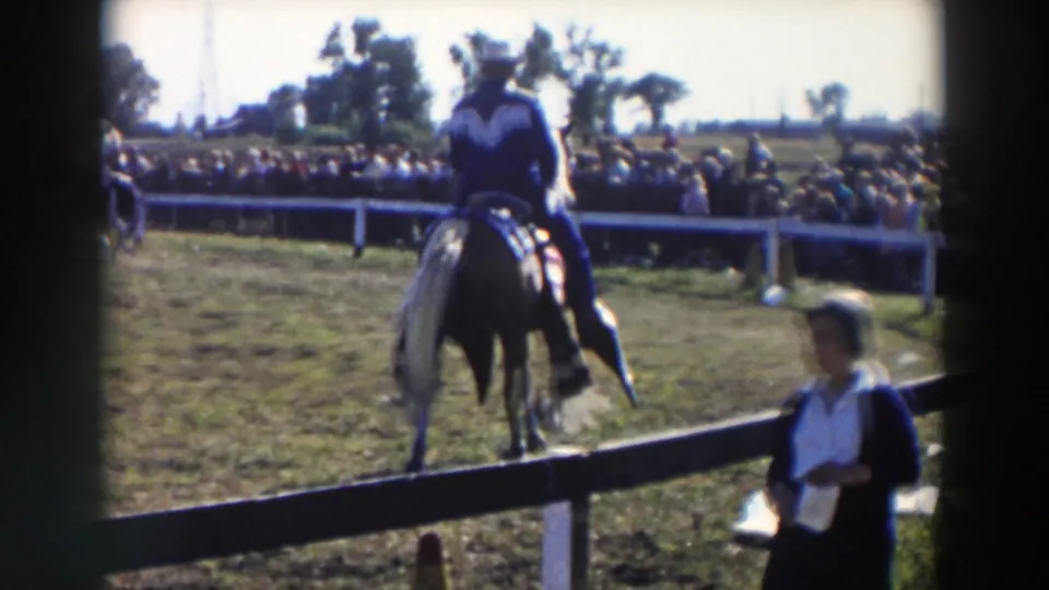 Rear view of the rider and horse in the Wheeling arena, with spectators beyond the fence and an adult woman near the foreground rail.