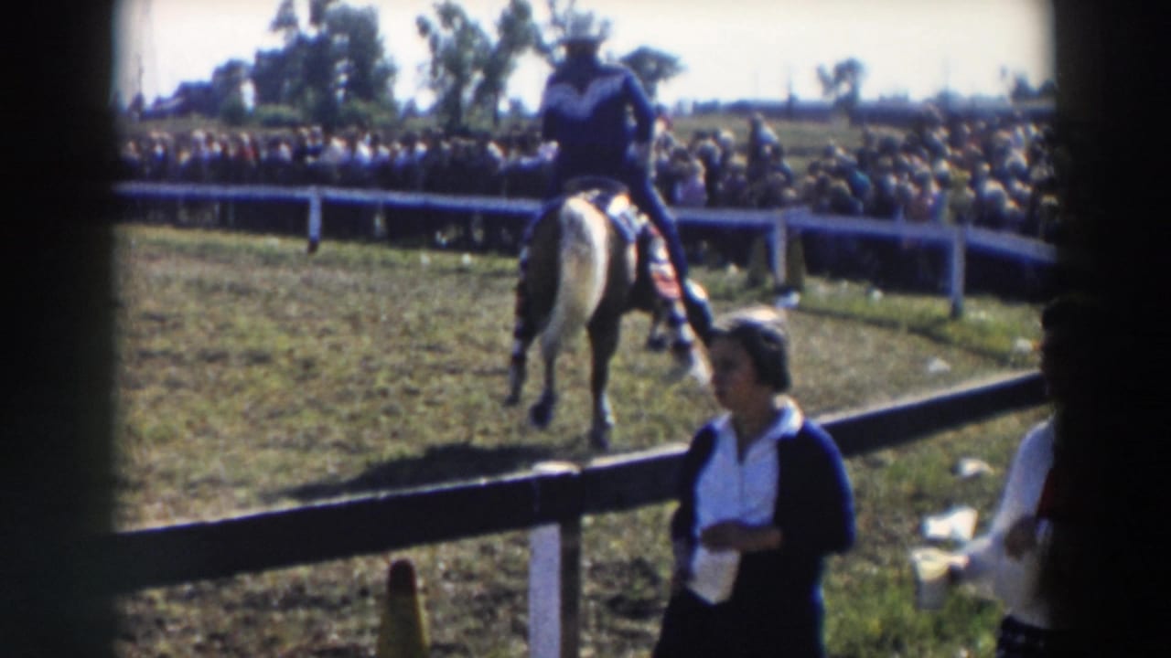 Rider on a light horse passes a white rail fence with spectators behind it.