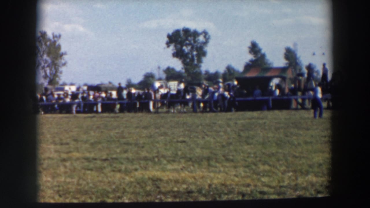 Wide view of a grassy riding ring with spectators near a small shelter.