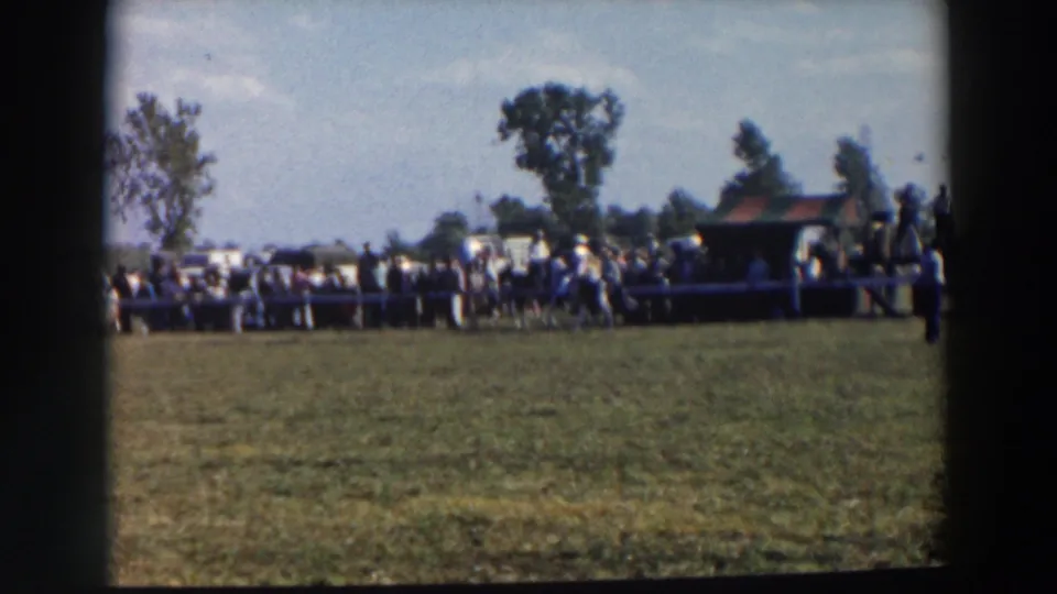 Home-movie view of spectators along the fenced riding field in Wheeling, Ohio, with trees and a small shelter in the background.