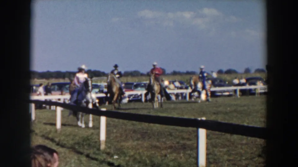 Several riders in hats guide horses around the fenced ring, with period cars and spectators visible beyond the rail.