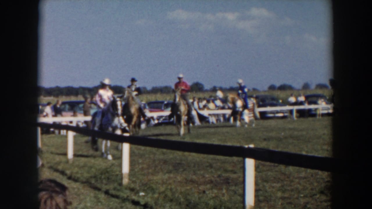 Mounted riders in cowboy hats move near a white fence on a grassy field.