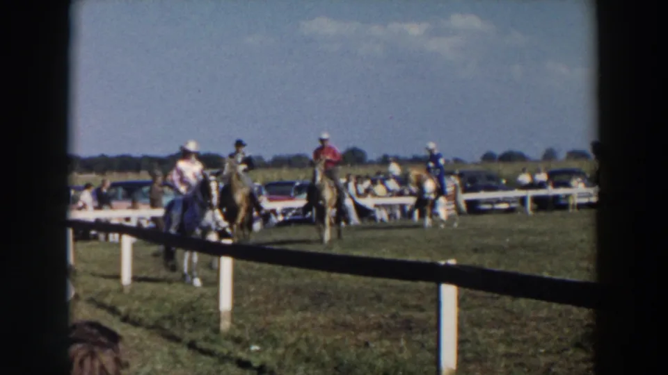 In 1957 Wheeling, Ohio footage, riders in cowboy hats guide horses past a white rail fence with parked cars behind them.