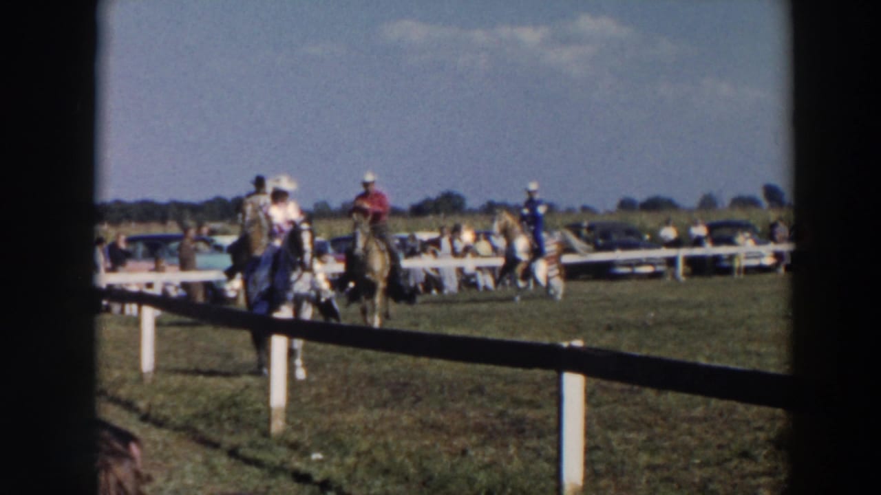 Several riders on horses gather near a fence with cars in the background.