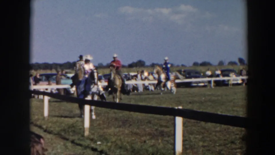 Riders in cowboy-style attire and hats appear on horseback at a Wheeling, Ohio public event, with mid-century cars beyond the fence.