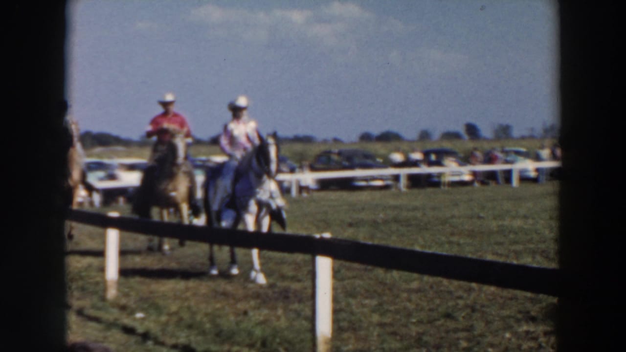 1957 Wheeling, Ohio home-movie frame showing two horseback riders in white hats near a white rail fence.