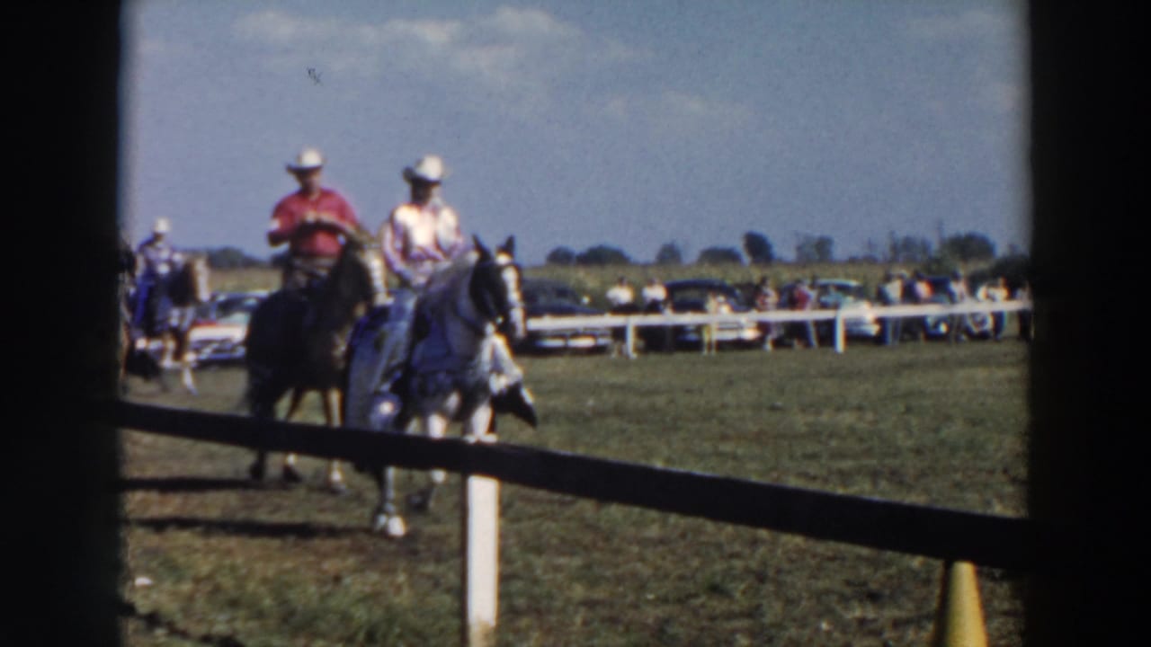 Horseback riders pass behind a dark foreground frame near a rail fence.
