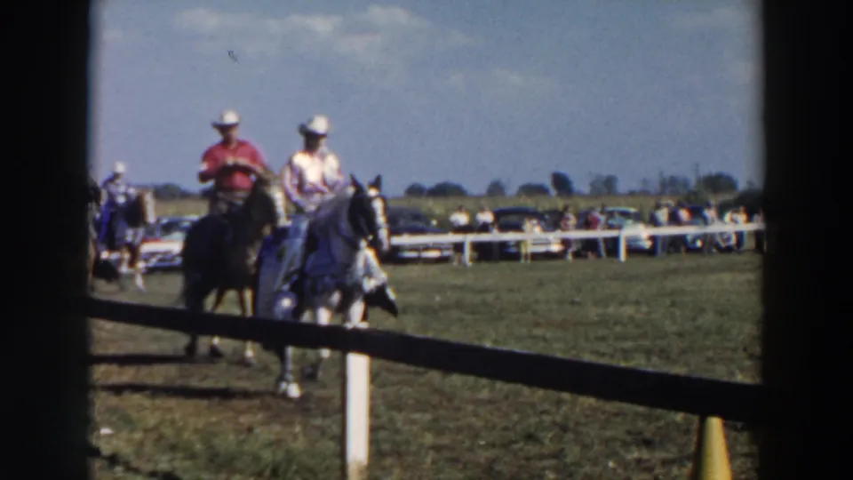 Two riders in white hats pass through a 1957 Wheeling, Ohio riding scene, with parked cars beyond the fence.