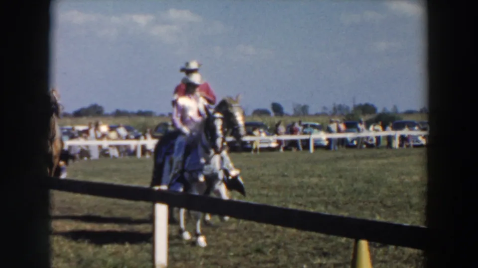 A white-hatted rider on a light-colored horse moves through a fenced 1957 Wheeling, Ohio riding field.