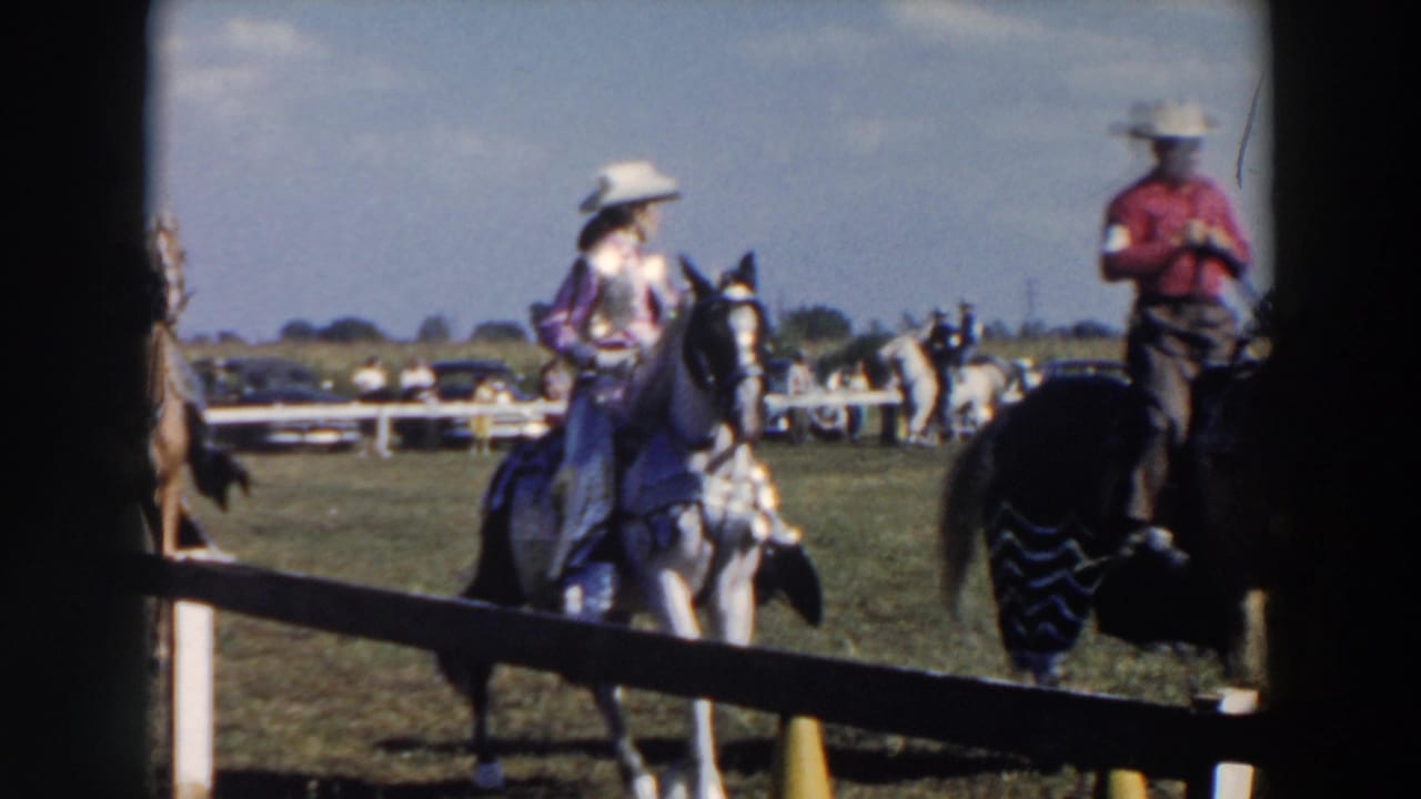 Mounted riders in cowboy hats circle a grassy horse-show ring behind a wooden rail.
