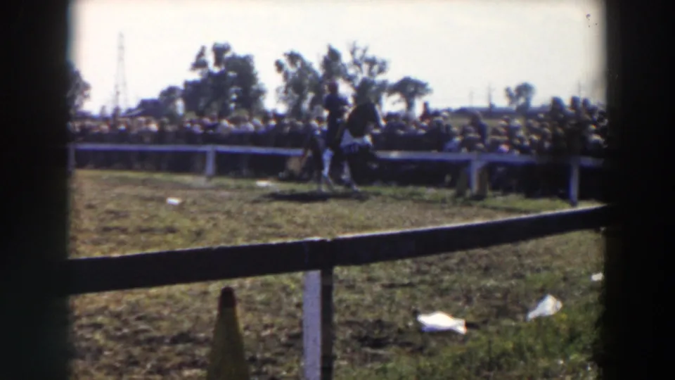 Wide 1957 home-movie frame of a grassy Wheeling, Ohio horse-show area seen through dark fence posts.