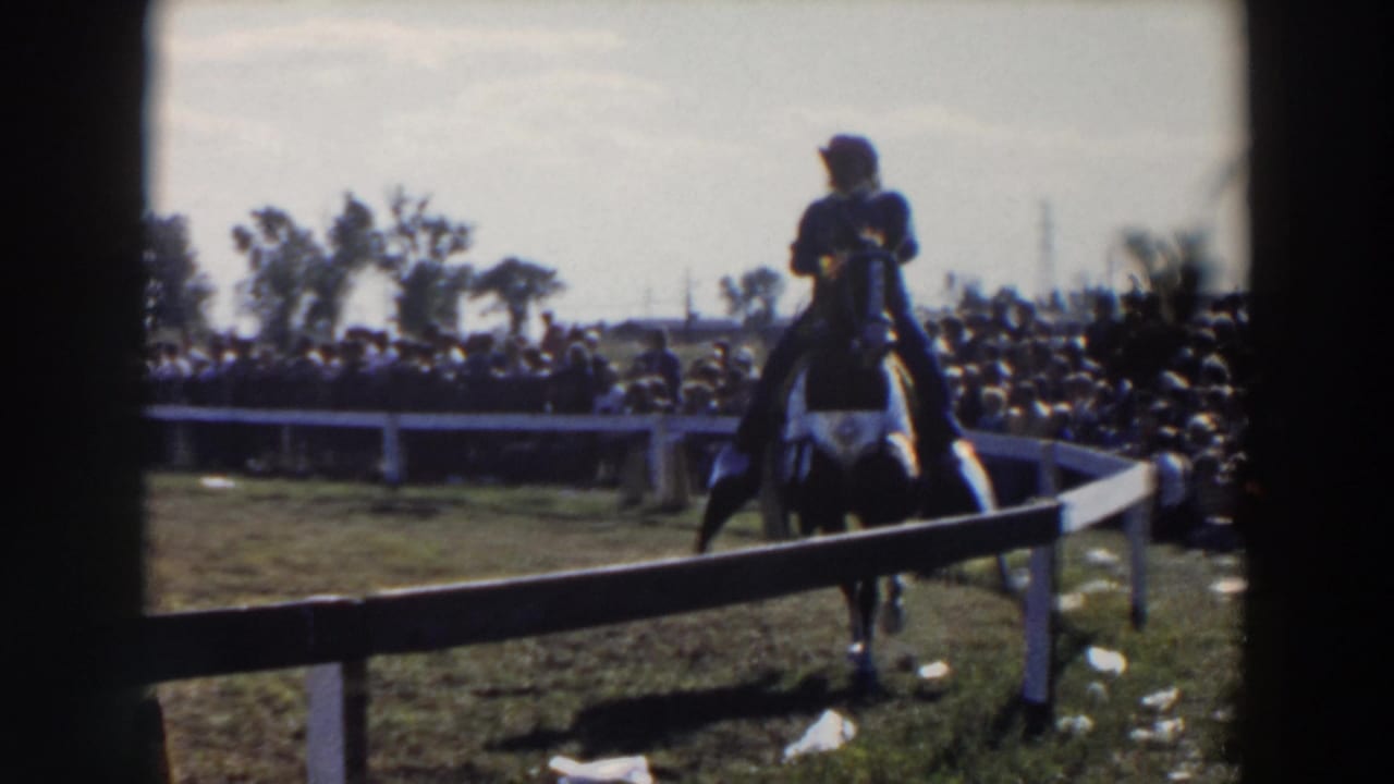 Mounted rider in a white hat approaches along a white rail fence before a crowd.