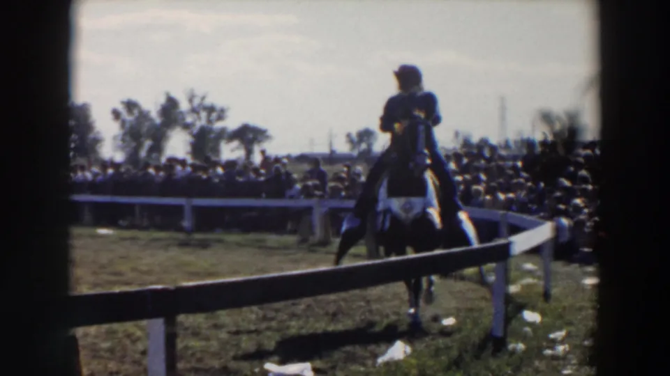 1957 Wheeling-area 8mm frame showing a rider in dark clothing and a white hat guiding a dark horse beside a white fence.