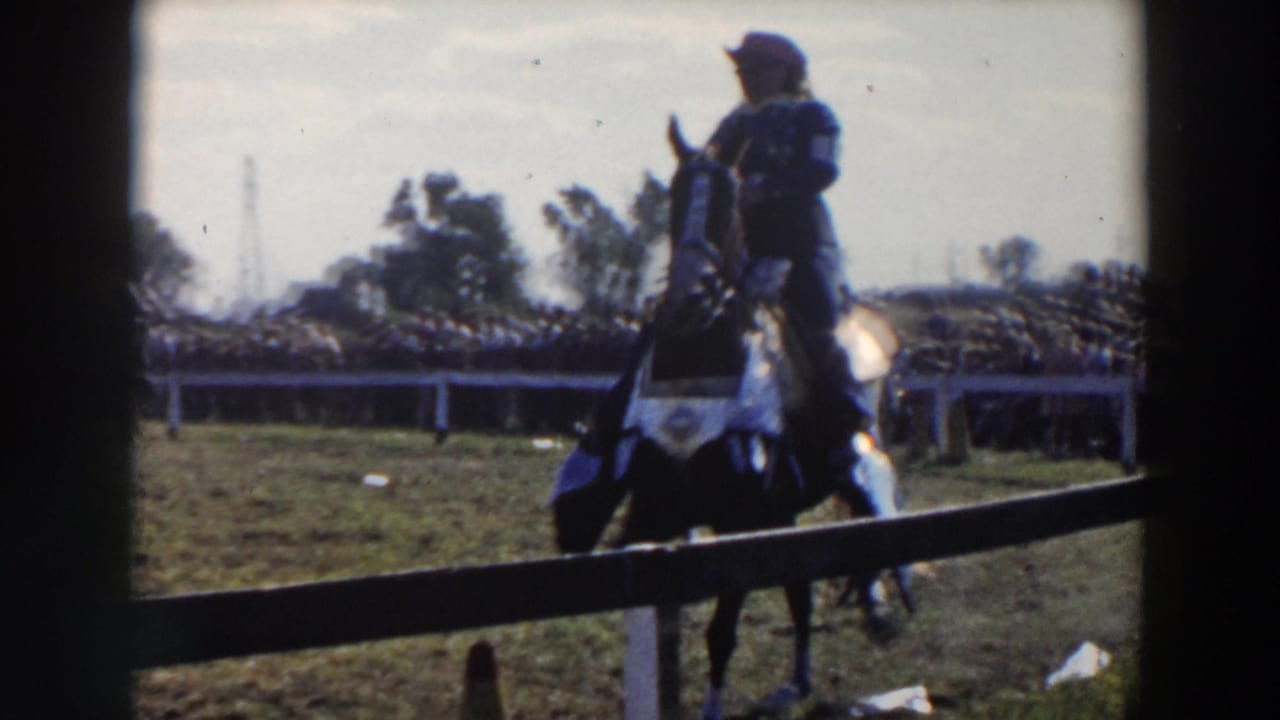 A dark horse and rider in a white hat pass near the camera at an outdoor equestrian event in the 1957 home-movie footage.