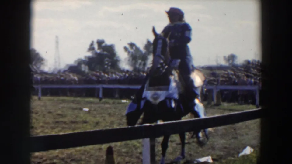 A dark horse and rider in a white hat pass near the camera at an outdoor equestrian event in the 1957 home-movie footage.