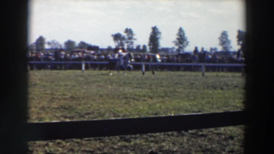 Wide 8mm view of the equestrian field in Wheeling-area footage, with a small rider and horse near the fence and spectators behind it.