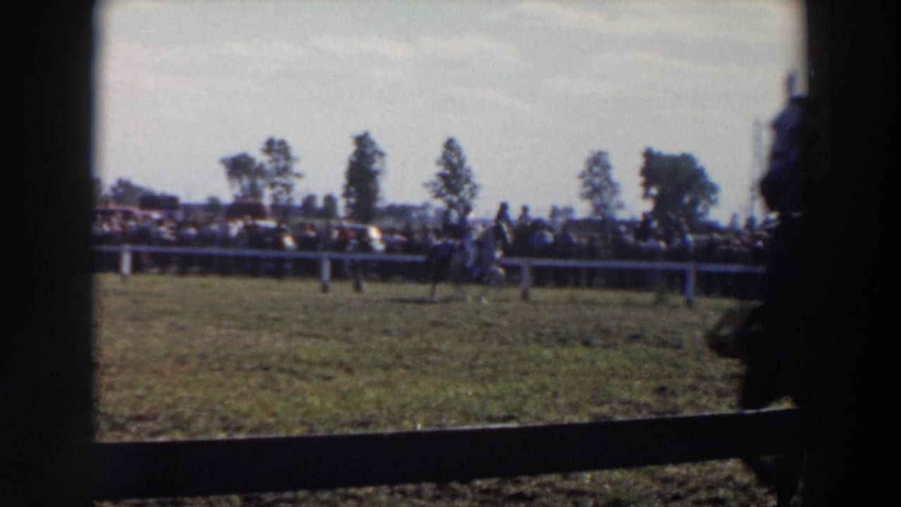 1957 Wheeling, Ohio home-movie frame showing horseback riders in a grassy enclosure with a crowd behind a white rail fence.