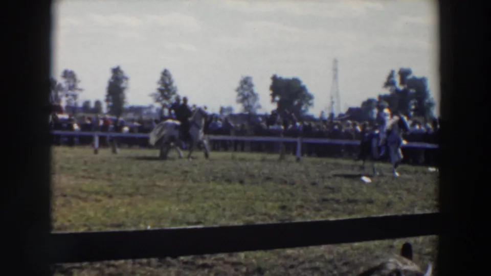 1957 Wheeling, Ohio home-movie frame showing multiple riders in the ring and a distant spectator line behind fencing.