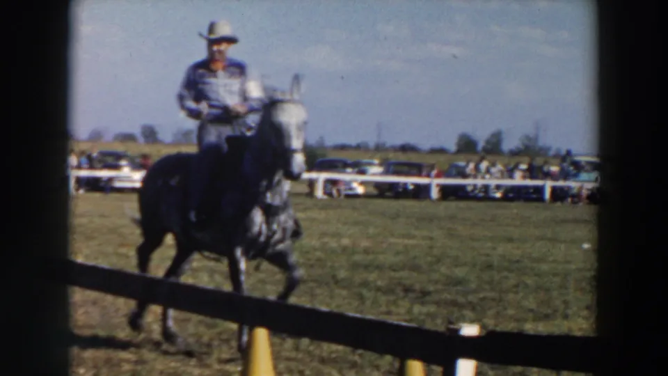 A rider wearing a cowboy hat approaches on a dark horse, with a white fence, yellow cones, and parked cars in the background.