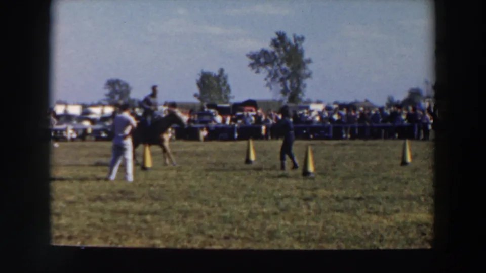 1957 8mm home-movie frame from Wheeling, Ohio, showing a rider guiding a dark horse near yellow cones before a spectator line.