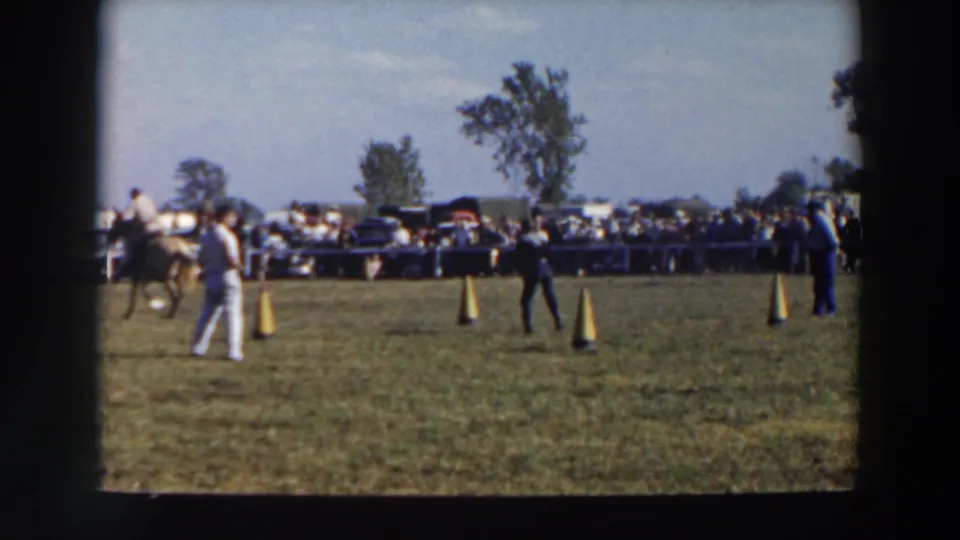 A horse moves out of frame at left while men remain on the cone-marked field and the spectator crowd looks on in the background.