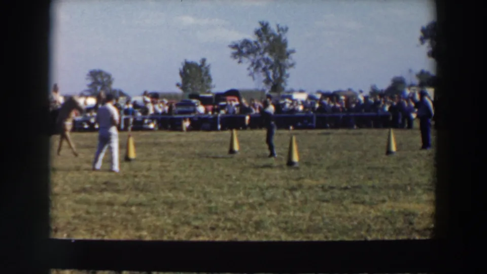 1957 Wheeling, Ohio home-movie frame showing a riding course on grass with cones, spectators behind a barrier, and cars beyond them.