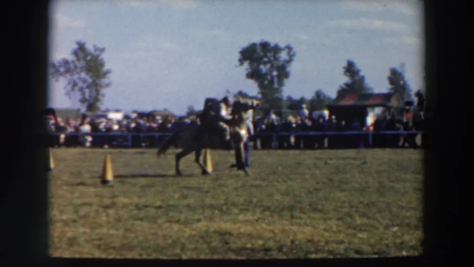 The rider guides the horse across the grassy course during the 1957 Wheeling, Ohio event, with a crowd and small building in the background.