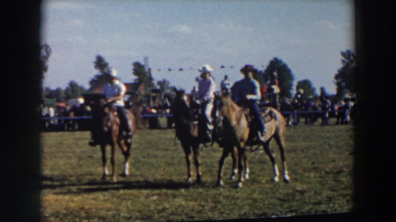 Mounted riders in cowboy hats pause on the grass during the Wheeling, Ohio horse event, with a spectator line and trees in the background.