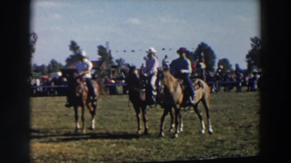Mounted riders in cowboy hats pause on the grass during the Wheeling, Ohio horse event, with a spectator line and trees in the background.