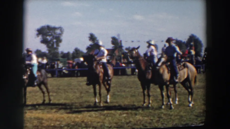 A group of mounted riders gathers in the competition field, with cowboy hats, horses, spectators, and trees visible in the home-movie frame.