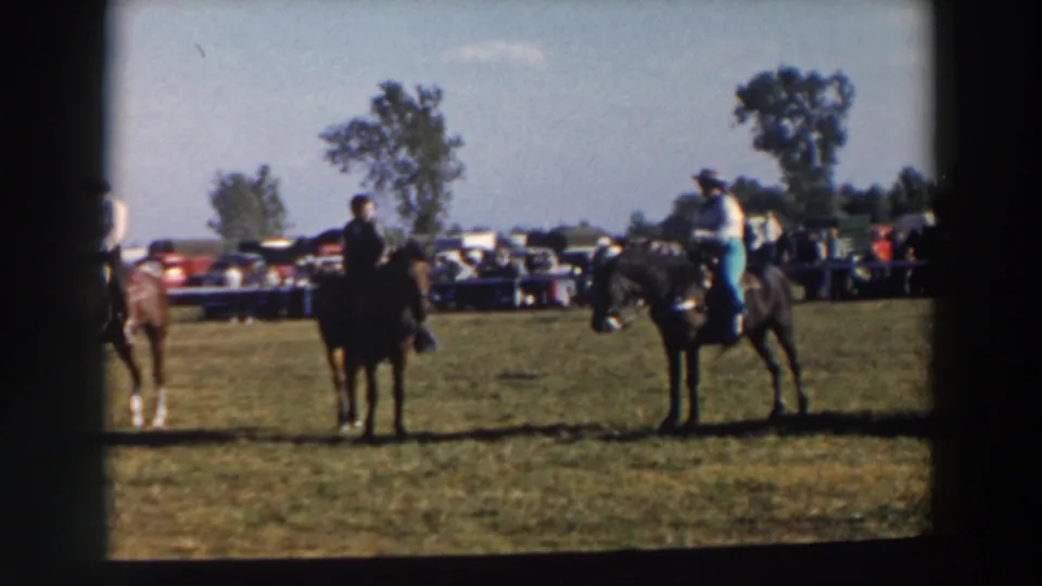 A row of horseback riders waits on grass at the Wheeling, Ohio event, with mid-century cars and onlookers behind them.