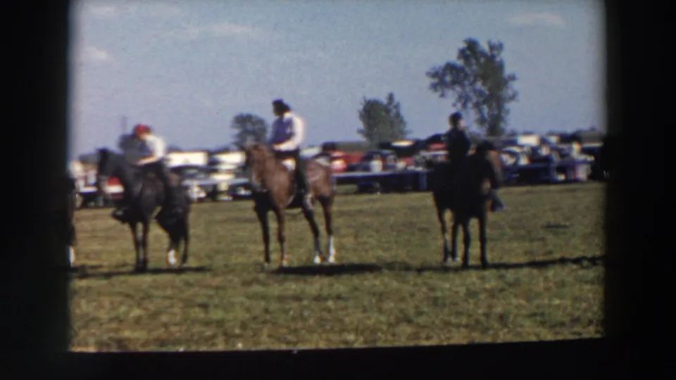 Several horseback riders wait across a grassy field in Wheeling, Ohio, with spectators and parked cars in the distance.