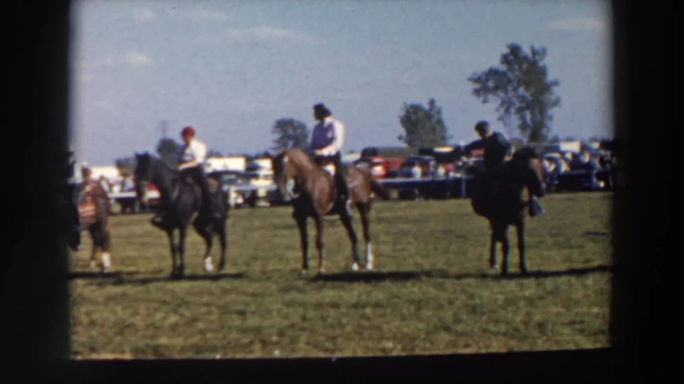 1957 Wheeling, Ohio home-movie frame showing riders on horses waiting in a grass field with spectators and parked cars in the background.